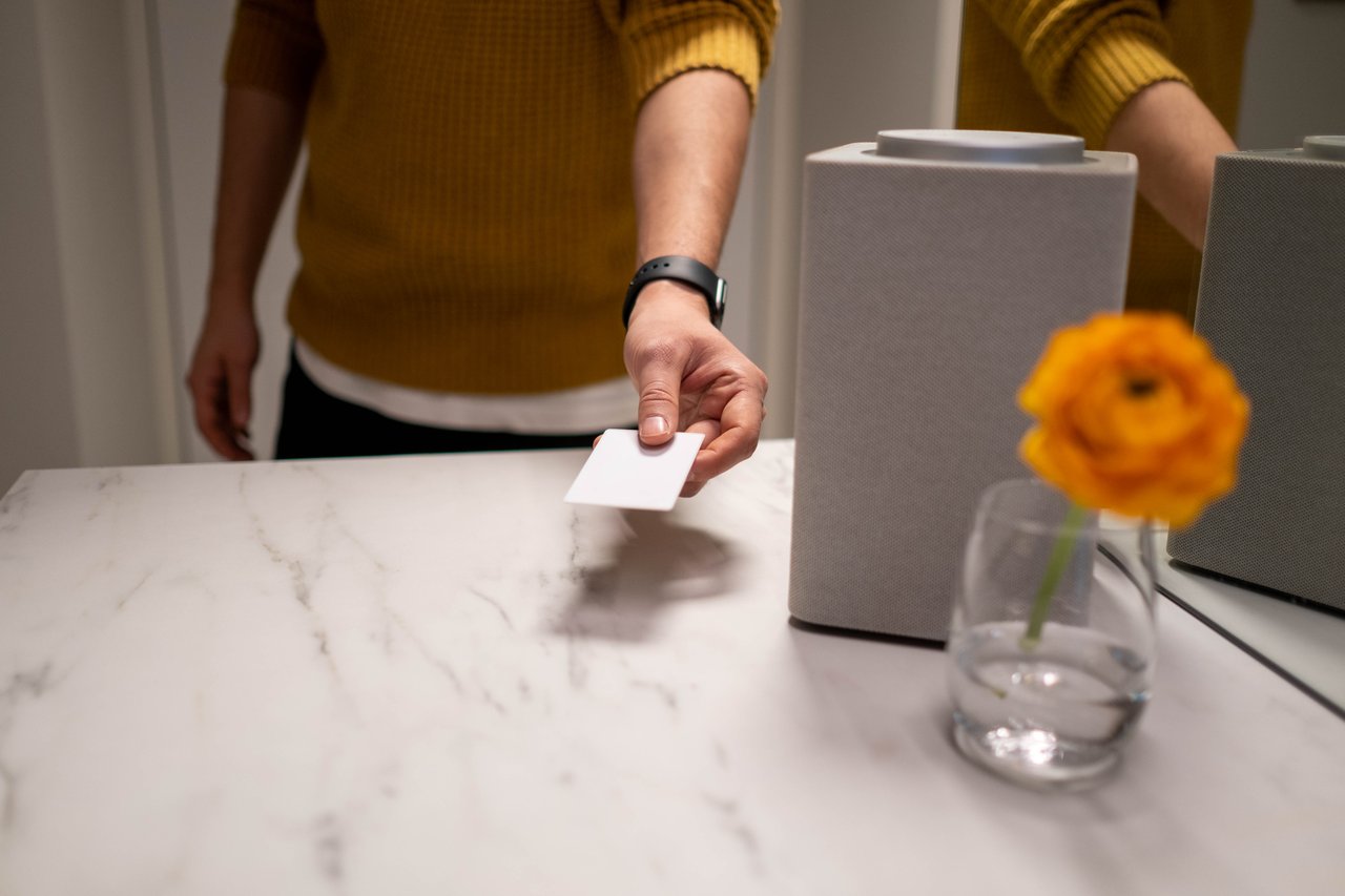 A man wearing a knitted sweater holds a card near a smart speaker on a marble table with a flower in glass.